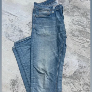 Ljusblå jeans från Nudie Jeans - Säljer ett par ljusblå jeans från Nudie Jeans med klassisk femficksdesign och snygga slitningar. Jeansen har raka ben och normal passform, tillverkade i mjuk bomull med Nudie-logga på bakfickan. Perfekta för en avslappnad stil. Byxorna är i storlek W31 L34