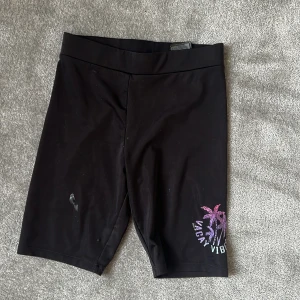 Svarta cykelshorts H&M Vacay Vibes - Svarta cykelshorts från H&M med tryck i lila och rosa av palmer och texten 'Vacay Vibes' på ena benet. Stretchigt material som sitter tight och är perfekta för sommaren eller träning. Midja i klassisk modell och längd till strax ovanför knät. Liten fläck i mitten av byxan, se bild 1