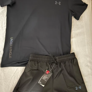 Svart Under Armour t-shirt med neontryck - Svart t-shirt från Under Armour med färgglada neontryck på bröstet, axeln, ärmen och nedtill. Klassisk rund hals och korta ärmar. Perfekt för dig som gillar sportig och modern stil. Materialet känns lätt och andas bra. (Finns även gråa) (Finns S, M och L)