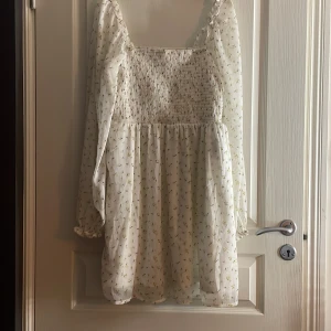 Vit blommig kortklänning Noisy May - Supersöt vit kortklänning från Noisy May med små blommor, smockad överdel och långa, luftiga ärmar. Klänningen har en flowy kjol och är perfekt för vår och sommar. Snygg fyrkantig urringning och diskreta volangdetaljer vid ärmslut.