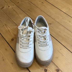 Vita sneakers med snörning - Säljer ett par stilrena vita sneakers med snörning. Skorna har en rund tå och en diskret randig detalj på sidan. 