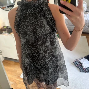 Svartvit mönstrad klänning med volangdetaljer - Säljer en svartvit kortklänning med höghalsad krage och volangdetaljer längs sidorna. Klänningen har ett abstrakt prickigt mönster och är ärmlös med lätt transparent material. 