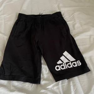 Svarta shorts från Adidas med logga - Svarta shorts från Adidas med vit logga tryckt på vänster ben. Modellen har resår i midjan med snörning och två sidofickor. Perfekta för träning eller avslappnade dagar. Tillverkade i mjukt material för extra komfort.
