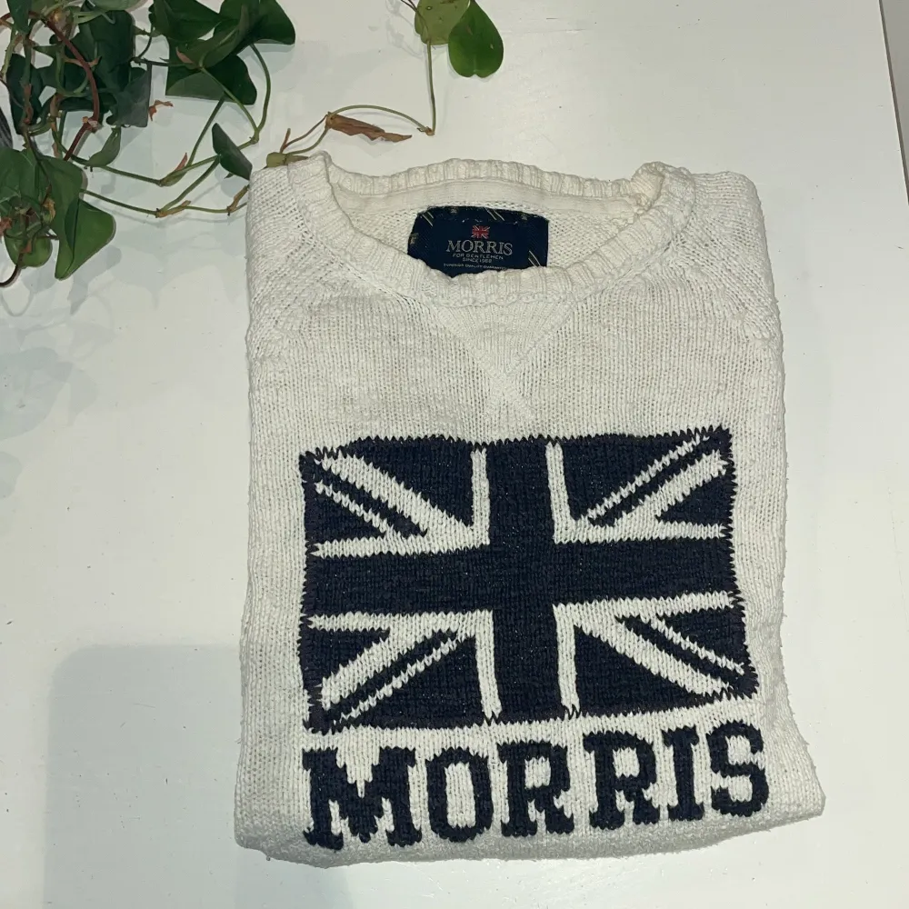 Vit stickad tröja från Morris med mörkblå brittisk flagga och texten 'Morris' framtill. Tröjan har rund halsringning och ribbade muddar vid ärmslut och nederkant. Perfekt för dig som gillar klassisk stil. Skriv vid minsta lilla fundering🙌. Neuletakit & Villapaidat.