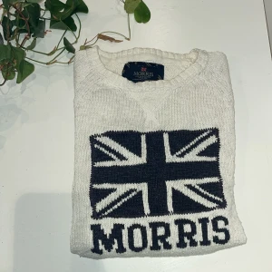 Stickad tröja från Morris - Vit stickad tröja från Morris med mörkblå brittisk flagga och texten 'Morris' framtill. Tröjan har rund halsringning och ribbade muddar vid ärmslut och nederkant. Perfekt för dig som gillar klassisk stil. Skriv vid minsta lilla fundering🙌