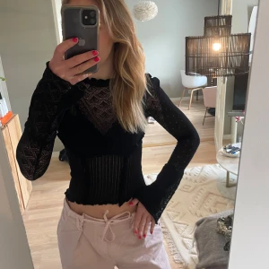 Svart croppad topp med spetsdetaljer - Säljer en svart croppad topp med långa ärmar och snygga spetsdetaljer. Tröjan har transparenta partier och volangdetaljer längs sömmarna. Perfekt för dig som vill ha en trendig och unik look. Den är något liten i storlek med ett litet hål på vänstra armen men som säkert går lätt att laga.