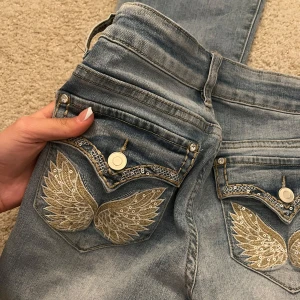 Blå jeans med broderade detaljer - Snygga blå jeans från madlady med unika broderade vingar på bakfickorna. Jeansen har en bootcut-stil och är dekorerade med spetsdetaljer vid fickorna. Perfekta för dig som vill ha något extra i garderoben! Storlek 32 💞använda en gång så inga defekter  
