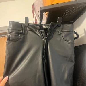 Svarta skinnbyxor från Zara - Snygga svarta skinnbyxor från Zara med dragkedja och knappdetaljer. Perfekta för en stilren look. Byxorna har en hög midja och en tight passform som framhäver figuren.