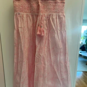 Ljusrosa långkjol  - Ljusrosa långkjol med smockad överdel och dekorativa tofsar. Perfekt för varma sommardagar med sitt lätta och luftiga material.