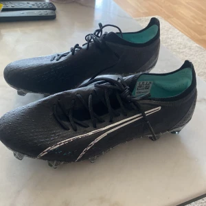 Puma ultra ultimate eclipse FG/AG - Säljer ett par svarta fotbollsskor från Puma med vit logga på sidan och mönstrad sula. Skorna har snörning och en låg profil, samt turkos insida. Perfekta för dig som vill ha stilrena och snabba skor på planen.