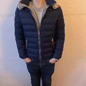 Mörkblå pufferjacka från Colmar - Säljer en snygg mörkblå pufferjacka från Colmar. Jackan har en dragkedja framtill och två praktiska fickor med dragkedjor. Den har en huva och är perfekt för kyligare dagar. Märkeslogga på ärmen ger en extra touch. Passar dig som är mellan 160-165 perfekt för skidor.