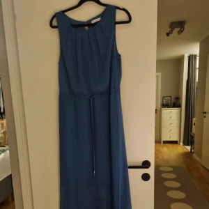 Petroleumblå ärmlös klänning - Säljer en fin petroleumblå ärmlös klänning med rund hals och band i midjan. Klänningen har en stilren design med veckade detaljer vid halsen och en knappstängning bak.