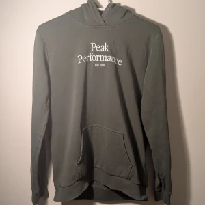 Grön hoodie från Peak Performance - Säljer en grön hoodie från Peak Performance med vit broderad logga på bröstet. Tröjan har känguruficka framtill och ribbade muddar vid ärmslut och nederkant. Perfekt för dig som gillar en klassisk och stilren look. Det är en hoodie i nästan nytt skick och inte alls sliten. Storlek 170                            Orginalpris 800kr