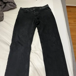 Mörkgrå jeans från Nudie Jeans strl:34/34  - Säljer ett par mörkgrå jeans från Nudie Jeans med klassisk femficksdesign och diskreta sömmar på bakfickorna. Jeansen har en avslappnad passform med raka ben, perfekta för en clean och stilren look.