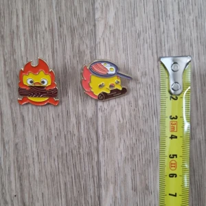 Färgglada pins med eldtema calcifer - Två färgglada pins med eldtema, calcifer från det levande slottet. Den ena föreställer en eld med ögon och ved, medan den andra har en eld med en stekpanna på huvudet. Perfekta som accessoarer för att ge en rolig touch till din outfit.