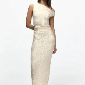 Krämfärgad oneshoulder långklänning från Pull&Bear - Säljer en jättefin krämfärgad långklänning från Pull&Bear med oneshoulder-design. Klänningen har en strukturerad, lätt skrynklig yta och figurnära passform som framhäver siluetten. Helt oanvänd med prislappen kvar! 