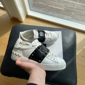 Valentino Garavani  - Säljer ett par vita sneakers från Valentino Garavani med en bred svart rem över vristen dekorerad med nitar. Skorna har en chunky vit sula och klassisk snörning. Perfekta för dig som vill ha en lyxig och edgy look.