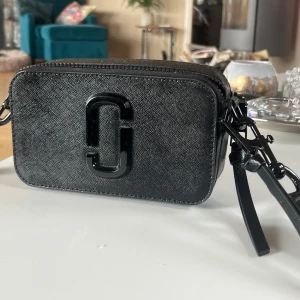 Svart väska från Marc Jacobs - Snygg svart axelväska från Marc Jacobs i modellen snapshot. Väskan har en dragkedja och en justerbar axelrem. Väskan är knappast använd men finns några repor på märket, men det syns endast på nära håll. Kan gå ner lite i pris vid smidig affär.