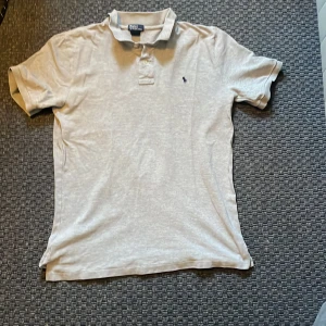 Beige kortärmad pikétröja från Polo Ralph Lauren - Säljer en stilren grå pikétröja från Polo Ralph Lauren. Tröjan är kortärmad och har en klassisk krage med knappar. Den är tillverkad i 100% bomull och har den ikoniska broderade logotypen på bröstet. Perfekt för en avslappnad och snygg look. Den här två mycket små hål i nacken som man inte tänker på men är värt att nämna. Annars inga fläckar eller liknande och knappt använd. Hör av dig vid minsta fundering.