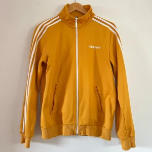 Gul träningsjacka från Adidas - Säljer en snygg gul träningsjacka från Adidas med vita ränder längs ärmarna och dragkedja framtill. Jackan har ribbade muddar och två fickor med dragkedja. Mycket bra skick.