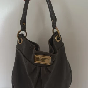  handväska från Marc by Marc Jacobs - Säljer en svart handväska från Marc by Marc Jacobs med guldfärgade metalldetaljer och en dekorativ metallplatta framtill. I använt skick (se bilder) därav priset