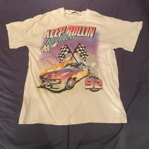 Vit t-shirt med biltryck från FB Sister - Cool vit t-shirt från FB Sister med stort tryck av en sportbil, eldflammor, rutiga flaggor och texten 'Keep Rollin Reckless' på framsidan. T-shirten har korta ärmar och klassisk passform. Perfekt för dig som gillar streetstyle och bilar.