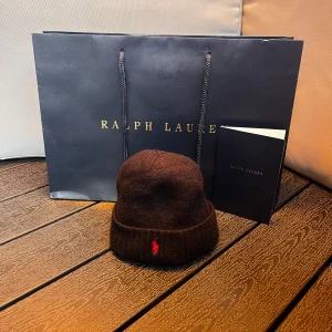 svart bebis mössa  - svart stickad bebis mössa med en röd broderad logga framtill. Mjuk och varm, perfekt för kyliga dagar. Tidlös design med uppvikt kant. för ungefär 1-2 åringar