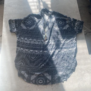 Mönstrad kortärmad topp från Lindex - Säljer en unik kortärmad topp från Lindex med ett iögonfallande geometriskt mönster i svart och grått. Toppen har en dragkedja framtill och en avslappnad passform. Perfekt för att ge din outfit en cool touch!