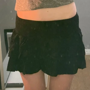 Svart volangkjol - Säljer en svart kortkjol med flera lager volanger och bred resår i midjan. Kjolen har en luftig och flowy känsla, perfekt för att skapa en söt och trendig look. Passar dig som gillar en feminin stil och vill ha något enkelt att matcha med olika toppar.