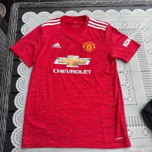 Röd Manchester United fotbollströja - Säljer en röd Manchester United fotbollströja från Adidas med korta ärmar. Tröjan har vita detaljer och klubbens emblem på bröstet. Perfekt för fans som vill visa sitt stöd på matchdagen!