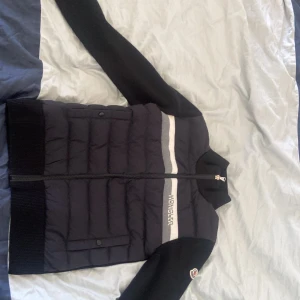 Svart dunjacka/cardigan från Moncler - Snygg svart dunjacka från Moncler med vita detaljer och dragkedja framtill.  Jackan har långa ärmar och en quiltad design som ger extra värme. Perfekt för kyligare dagar.
