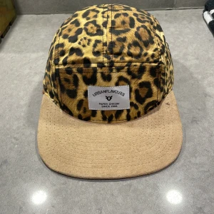 Leopardmönstrad keps från Urban Flavours - 5 panel - Snygg leopardmönstrad keps från Urban Flavours med beige skärm. Kepsen har justerbart spänne baktill för perfekt passform. Perfekt för att ge din outfit en djärv touch.