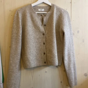 Beige stickad kofta från JDY - Mysig beige stickad kofta från JDY med rund halsringning och knappar framtill. Tröjan har lång ärm och en klassisk, enkel design som passar till mycket. Perfekt för lager-på-lager och chill dagar.