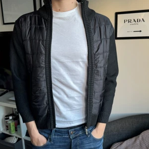 Svart cardigan jacka med stickade ärmar - Snygg svart cardigan jacka med stickade ärmar och rygg samt vadderad framsida. Jackan har dragkedja och ribbade muddar. Perfekt för lager-på-lager och en stilren look.