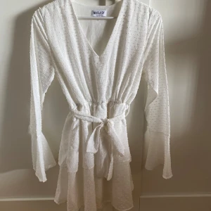 Klänning - Superfin vit kortklänning från Madlady med v-ringning, volangdetaljer och knytband i midjan. Klänningen har långärmade ärmar med volang vid ärmslut och ett prickigt mönster i tyget.