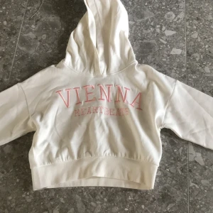 Vit hoodie från Lindex med rosa text - Säljer en vit hoodie från Lindex i storlek 146/152. Tröjan har huva och texten 'VIENNA HEARTBEATS' i rosa på ryggen. Perfekt för dig som gillar en enkel men snygg stil.