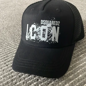 Icon Dsquared2 keps - Säljer en svart keps från Dsquared2 med stor vit text 'ICON' framtill och 'Born in Canada' baktill. Kepsen har mesh på sidorna och bak, samt justerbar snapback. Perfekt för en cool och avslappnad stil. Helt ny. 