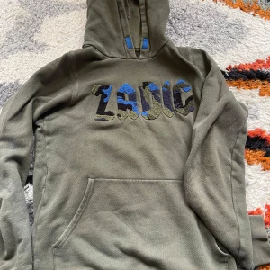 Olivgrön hoodie från Zadig & Voltaire - Säljer en olivgrön hoodie från Zadig & Voltaire med broderad logga i blått, svart och grönt framtill. Tröjan har huva, känguruficka och mjukt material. Perfekt för en avslappnad stil. Har inte sett en liknande innan så gissar på att den är ganska ovanlig.