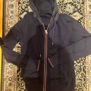 Mörkblå cardigan med dragkedja från Moncler - Säljer en mörkblå hoodie från Moncler med dragkedja och huva. Tröjan har ribbade muddar, fickor framtill och snygga rödvita detaljer längs dragkedjan. Perfekt för dig som gillar stilrena och exklusiva plagg. Storleken inuti är xl men den passar M-L