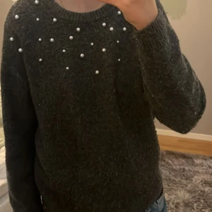 Mörkgrå stickad tröja med pärlor från H&M - Säljer en mörkgrå stickad tröja från H&M med dekorativa pärlor framtill vid halsringningen. Tröjan har lång ärm och rund hals, perfekt för en mysig och stilren look. 