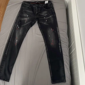 Svarta jeans med färgstänk från Dsquared2 - Säljer ett par svarta jeans från Dsquared2 med coola färgstänk och slitningar. Jeansen har fem fickor, röd söm på insidan och är tillverkade i bomull. Passar dig som vill sticka ut med en unik stil.