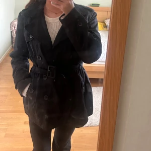 Svart trenchcoat med bälte - Säljer en svart trenchcoat med bälte i midjan och dubbelknäppning. Jackan har klassisk krage, långa ärmar och fickor på sidorna. Perfekt för dig som vill ha en stilren och tidlös look.