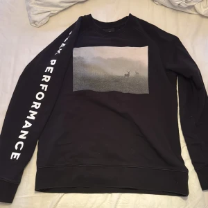 Svart sweatshirt från Peak Performance med tryck - Svart sweatshirt från Peak Performance med landskapsmotiv och hjort på bröstet. Texten 'Peak Performance' står längs ena ärmen. Klassisk rund hals och långa ärmar. Köpt ny och aldrig använd. 