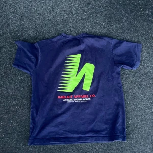 Mörkblå t-shirt från Harlaut Apparel - Säljer en mörkblå t-shirt från Harlaut Apparel aldrig använd. med grön logga på bröstet och stor grafisk print på ryggen. Perfekt för en sportig och avslappnad stil.