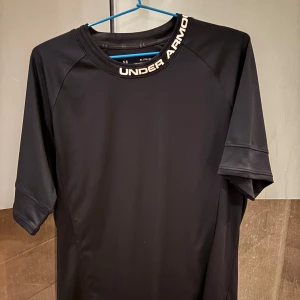 Svart träningst-shirt från Under Armour - Säljer en svart kortärmad träningst-shirt från Under Armour med vit logga och text runt halsen. Tillverkad i syntetmaterial och har en fitted passform, perfekt för träning eller sportaktiviteter.