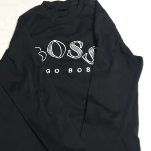 Svart långärmad tröja från Hugo Boss - Säljer en svart långärmad tröja från Hugo Boss med stor vit BOSS-logga framtill. Tröjan har rund halsringning och är i ett mjukt material, perfekt för en stilren och enkel look. Det finns håll men de (syns inte )