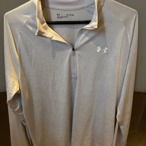 Vit långärmad träningströja från Under Armour - Säljer en vit långärmad träningströja från Under Armour med halv dragkedja framtill och diskret logga på bröstet. Perfekt för träning eller löpning, tillverkad i ett lätt och syntetiskt material som andas.
