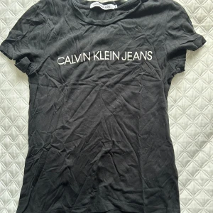 Svart t-shirt från Calvin Klein Jeans - Säljer en klassisk svart t-shirt från Calvin Klein Jeans med vit logga på bröstet. Perfekt för en stilren och enkel look. T-shirten är kortärmad och gjord i mjuk bomull.