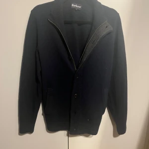 Cardigan från Barbour - Säljer en mörkblå stickad kofta/cardigan från Barbour med dragkedja och knappar framtill. Som ny, inga defekter. Den har bruna mockadetaljer på armbågarna. 