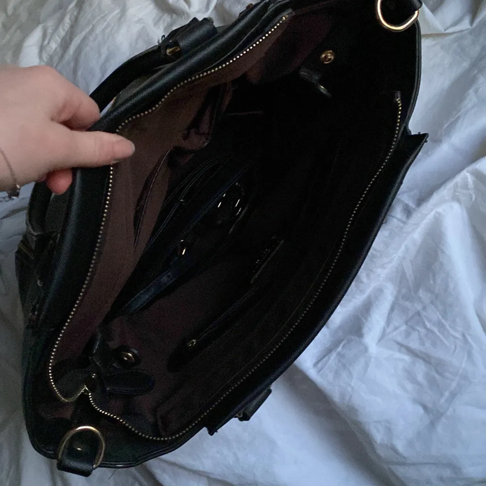 Snygg svart handväska från Michael Kors med guldfärgade detaljer och dragkedjor. Väskan har ett stort huvudfack och flera mindre fack inuti för enkel organisering. Perfekt för att bära med sig alla nödvändigheter med stil.. Laukut & Käsilaukut.
